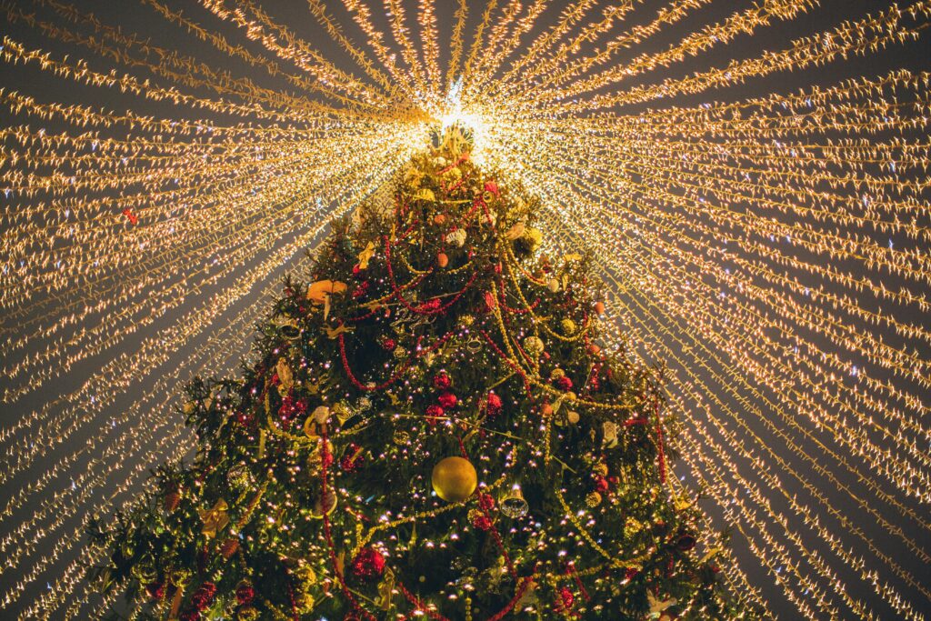 A large, beautifully decorated Christmas tree covered in ornaments, garlands, and glowing lights, with hundreds of golden string lights radiating outward from the top, creating a starburst effect against the night sky.