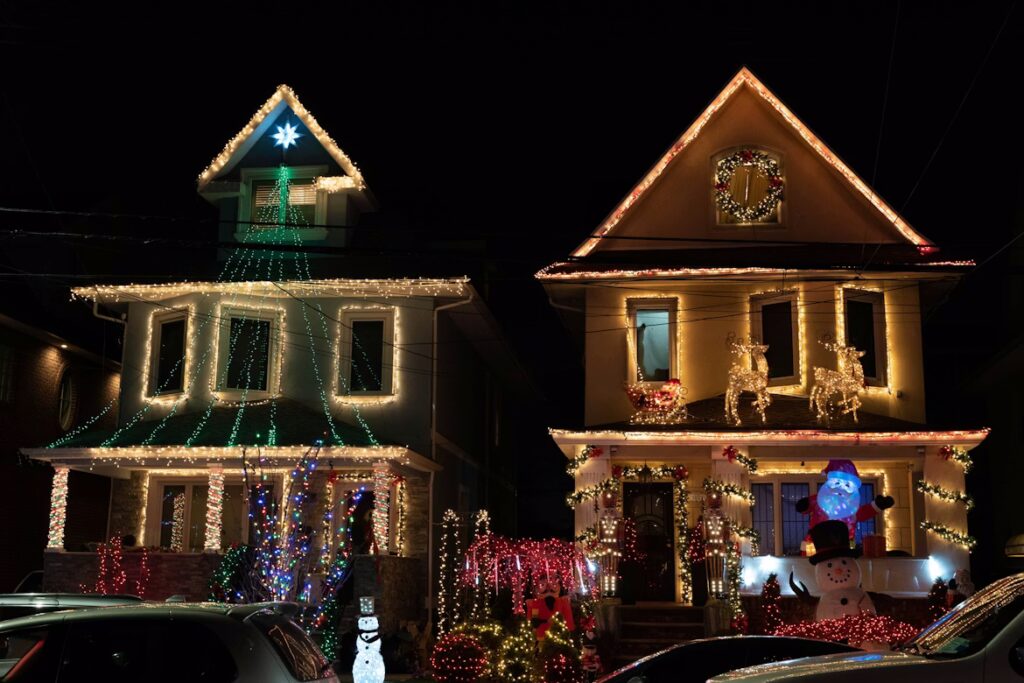 Two neighboring houses fully decorated with colorful Christmas lights at night. The left house features a tree-shaped green light display and a glowing star on the roof, while the right house displays reindeer, Santa Claus inflatables, and garlands with warm white lights.