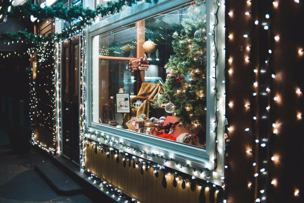 Festive shop window decorated with Christmas lights, garland, and a small Christmas tree. Presents and a cozy blanket are displayed inside, hinting at holiday gifts. String lights highlight the building's exterior.