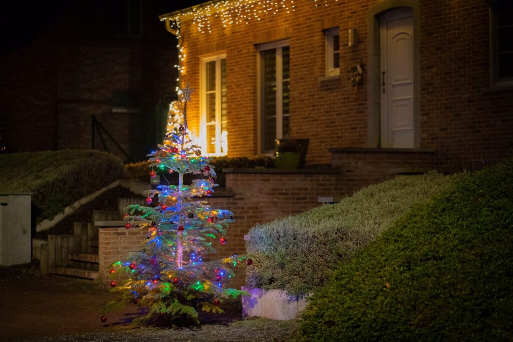 Outdoor Christmas tree adorned with colorful lights and ornaments, standing in front of a brick house with holiday lights. A festive and inviting scene, perfect for showcasing Christmas light design ideas.