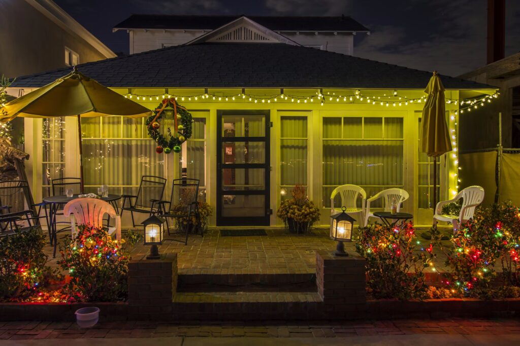 Festive home exterior at night, decorated with Christmas lights and a wreath. Warm yellow string lights illuminate the porch, bushes glow with colorful bulbs, creating a cozy holiday scene.